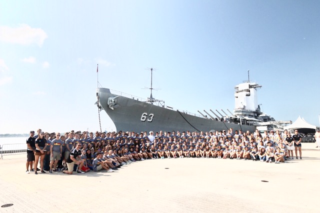 Band, choir, and dance students stand in front of a naval ship