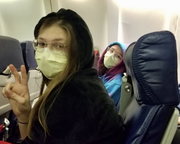 A student and her friend sit side-by-side on an airplane wearing surgical masks. 