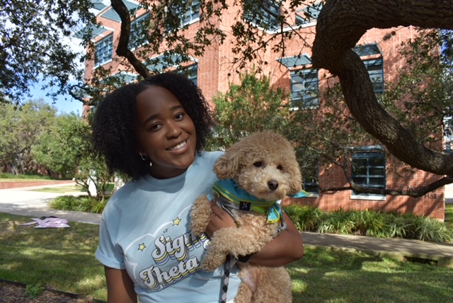 Kelli Steward stands outside the Sigma Theta Tau house at Trinity, wearing her Sigma Theta Tau shirt and holding a dog